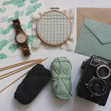 A watch, cross stitch piece, envelopes, yarn and knitting needles, and a camera on a table.