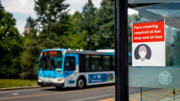 A TCAT bus shelter with a sign reading "Face covering required at bus stop and on bus" with a TCAT bus in the background.