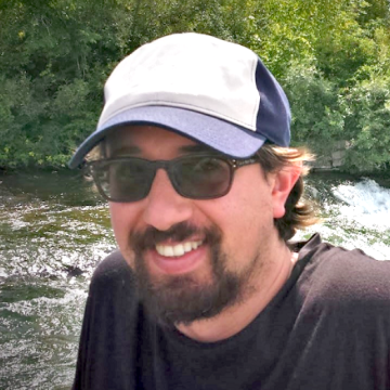 Sebastian outside by a river in hat and glasses