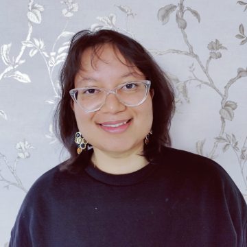 Shagun in glasses and earrings, smiling in front of ornate wallpaper