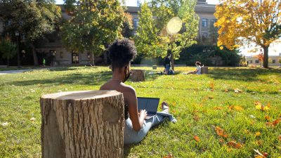 A student sits with her back against a tree stump on the Ag Quad on Cornell's Ithaca campus