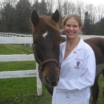 Erica Secor, wearing a white lab coat with a Cornell Veterinary Medicine logo, stands with a horse