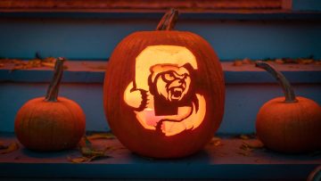 A lit jack-o-lantern carved with Touchdown coming through the Cornell C on a step with smaller, uncarved pumpkins on either side