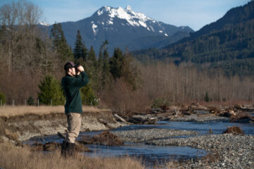 Ethan Duvall looks through binoculars with water and mountains in the background
