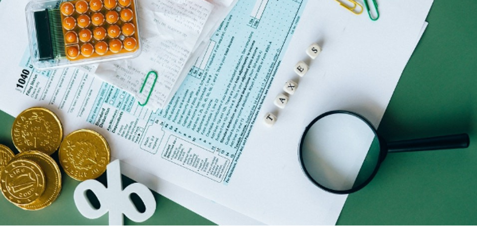 Green surface covered with papers, a calculator, gold coins, a magnifying glass and letters spelling "taxes"