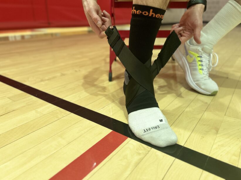 An athlete stands on a basketball court putting on the rheohero ankle support