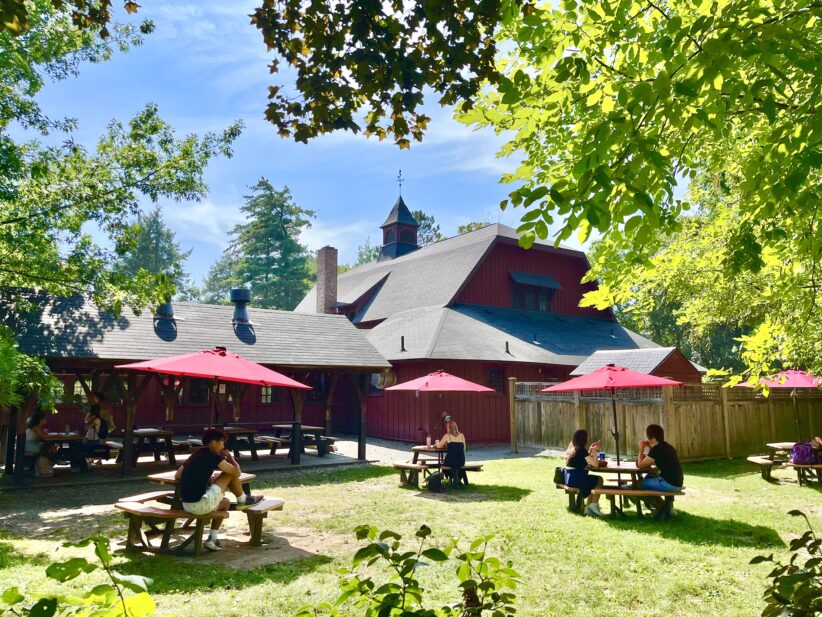 Picnic tables with red umbrellas in the grass outside a large barn