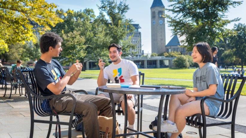 Three students sit at an outside table with McGraw Tower in the background