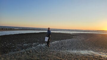 Associate Professor Matt Hare stands on the sand in front of a body of water