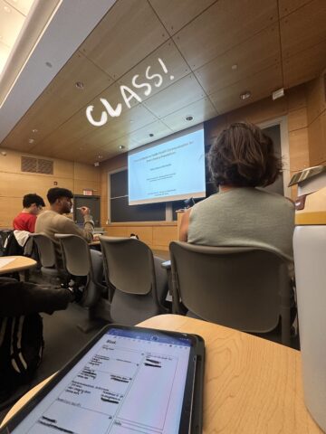 A tablet is open on a desk near the back of a classroom with students ahead looking at a large screen