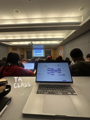 A laptop is open on a desk behind rows of students looking at a large screen at the front of the classroom