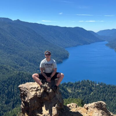 John Miller sits atop a rock with mountains and a lake stretched out behind him