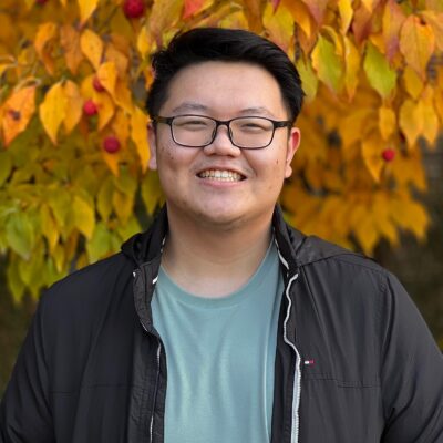 Jason Wu stands outside in front of a tree with bright fall foliage