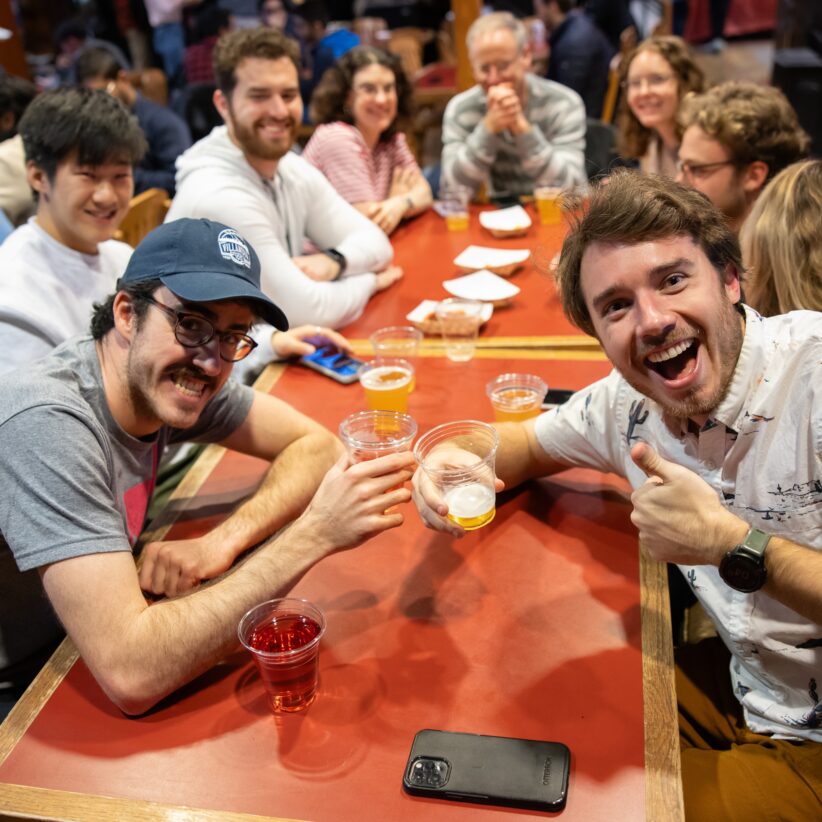 Students sit around a table at the Big Red Barn during a TGIF event, holding beer and smiling at the camera