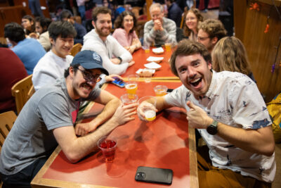 Students sit around a table at the Big Red Barn during a TGIF event, holding beer and smiling at the camera