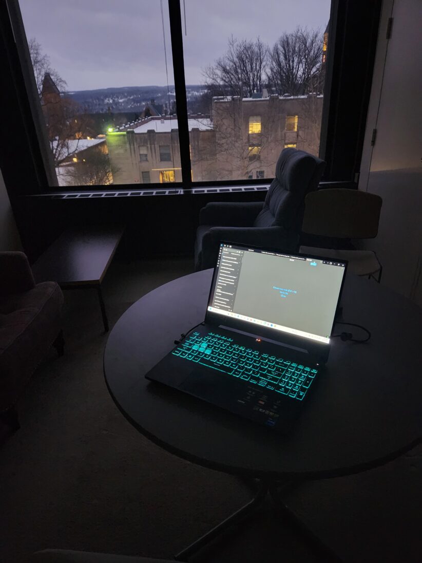 A laptop sits open on a table while a dusky view of campus is seen through the window behind it
