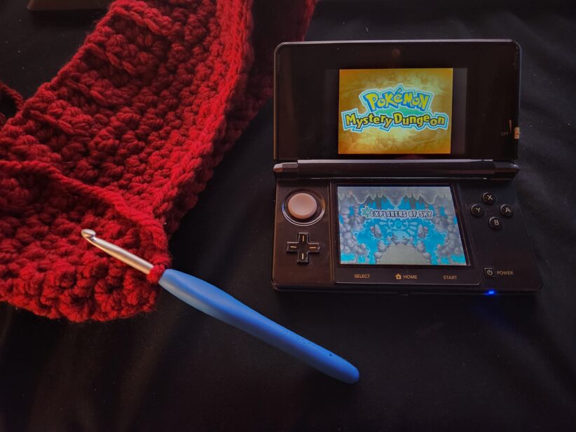 A crochet hook and project sit on a table next to a Nintendo 3DS open with the Pokémon Mystery Dungeon Explorers of Sky game on