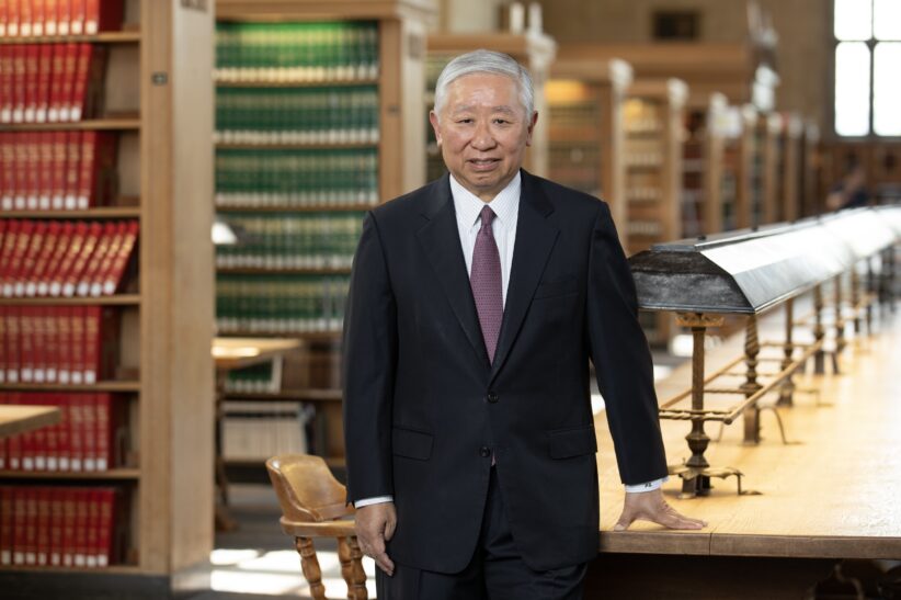 Jonathan Zhu stands in the Cornel Law Library with his hand on a long table and rows of shelves behind him