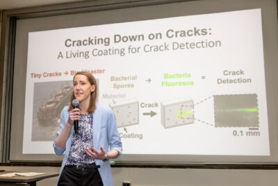 Ellen van Wijngaarden stands in front of her slide, microphone in hand, giving her three minute presentation.