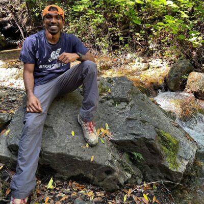 Deepak Varuvel Dennison sits atop a rock next to a small waterfall with natural greenery behind him