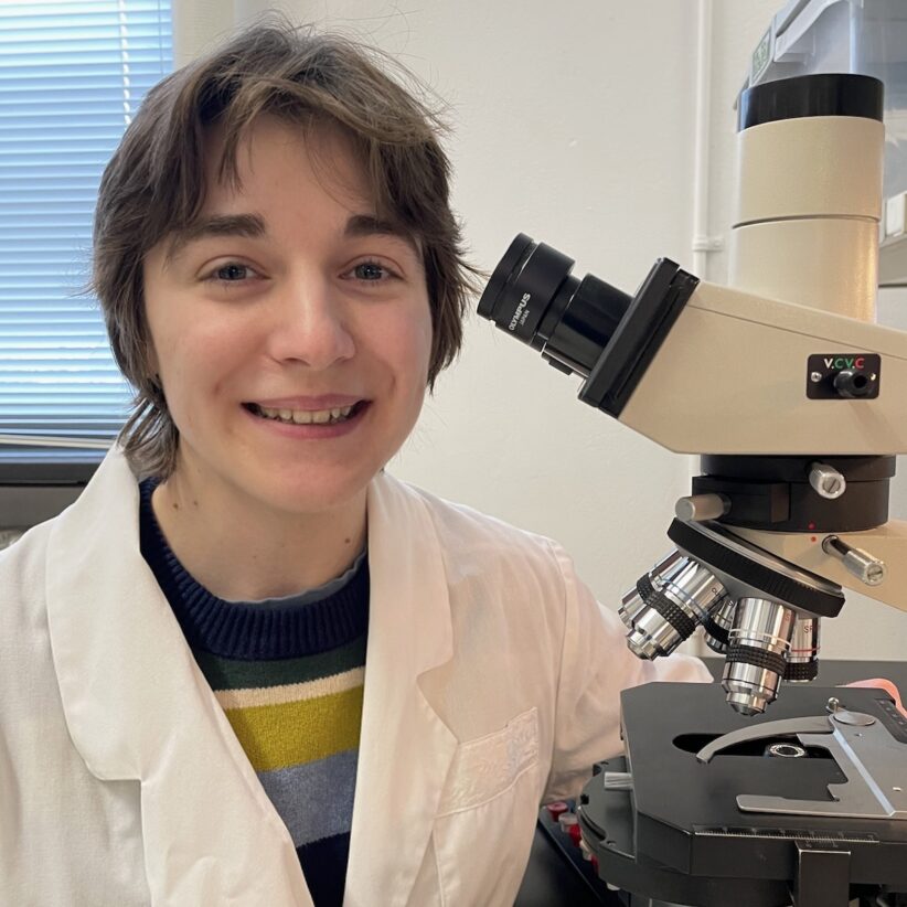 Delia Tota wears a white lab coat and sits next to a microscope, smiling at the camera
