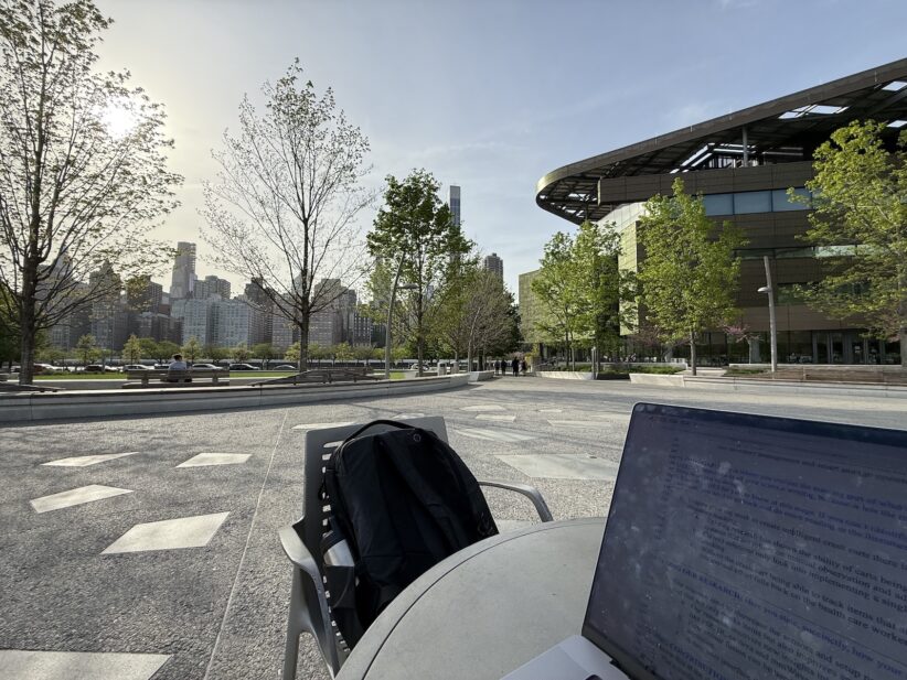 An open laptop sits on a table with a backpack on a chair with Cornell Tech visible in the background