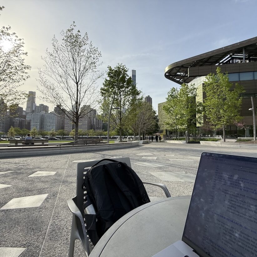 An open laptop sits on a table with a backpack on a chair with Cornell Tech visible in the background