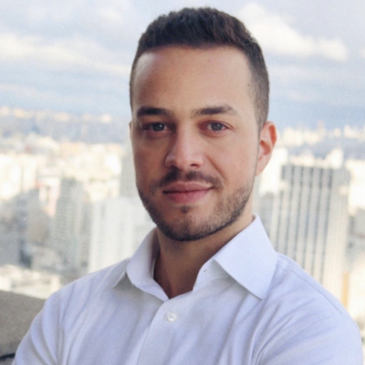 Manoel Pereira Neto stands in front of a window where tall city buildings stretch out behind him