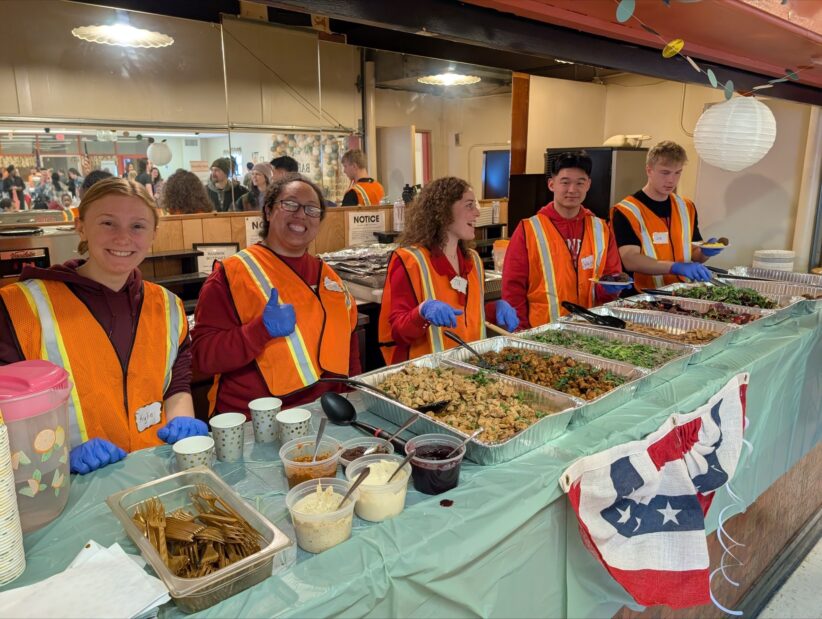 A line of student volunteers wearing bright colored safety vests stand behind a serving line of food trays