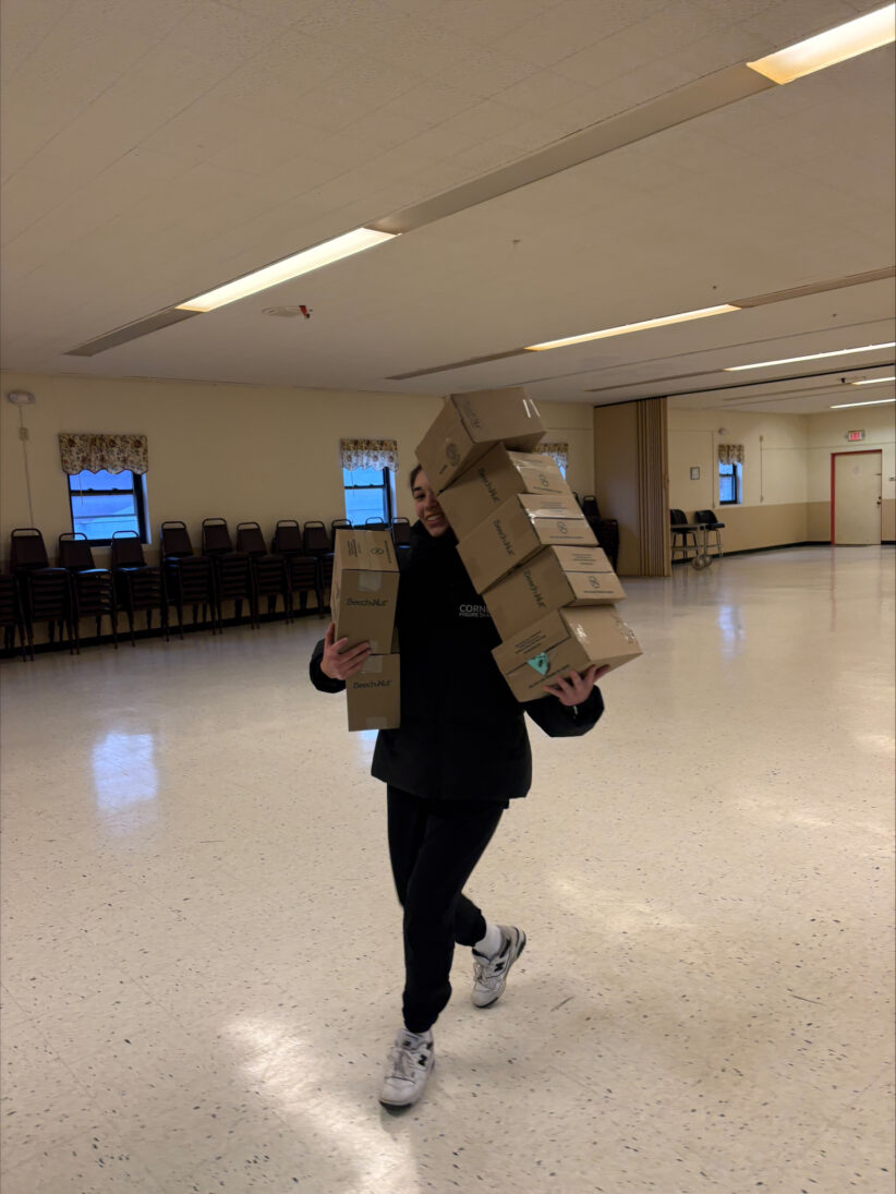 A student walks across a room carrying large stacks of boxes containing baby snacks