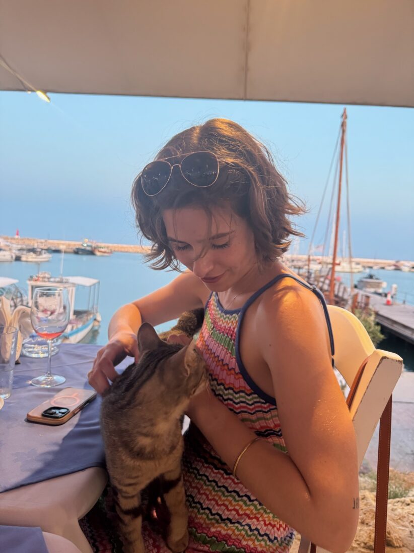 A cat stands on Anna Port's lap as she sits at a dining table with boats in a harbor seen out the window behind her