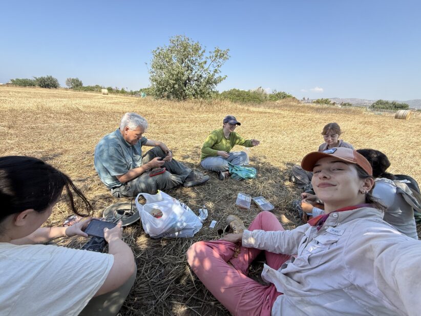 Anna Port sits on the ground near her archaeological site with other researchers during a snack break