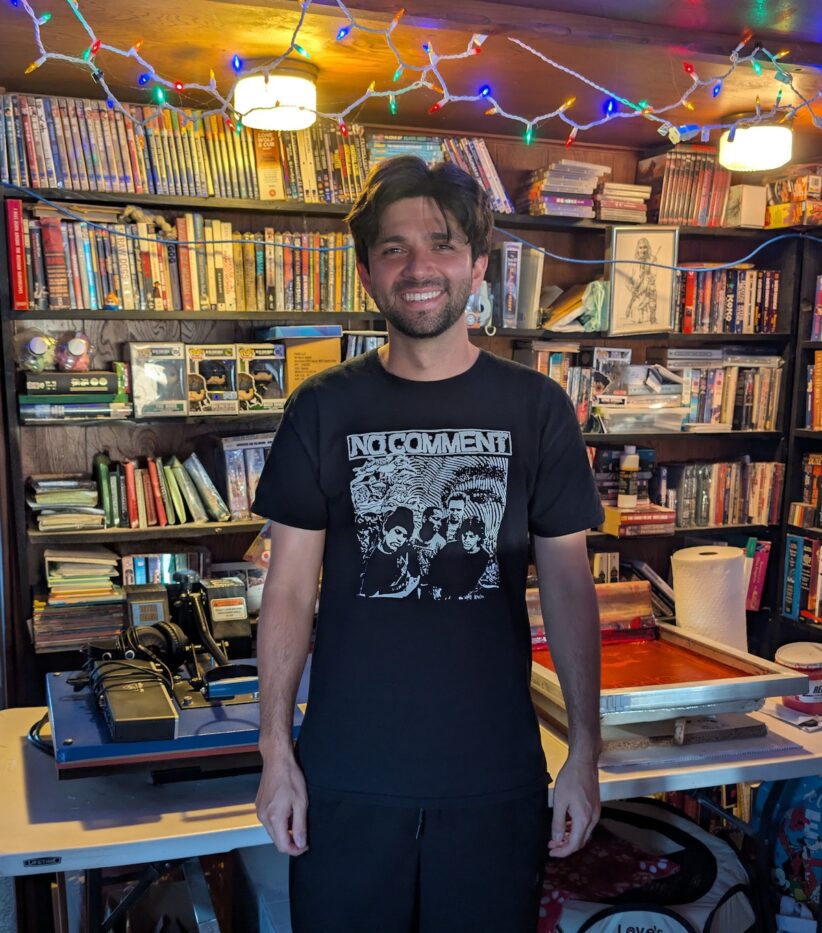 Michael Millenheft stands in front of a table with screenprinting tools on it with bookshelves filled with books and other items in the background