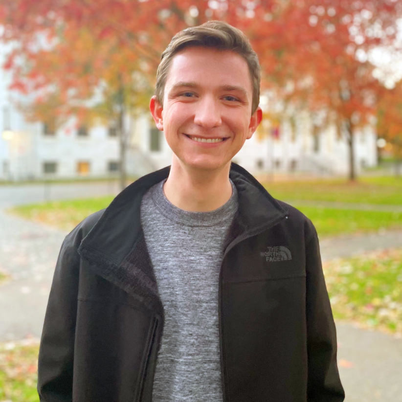 Nicholas Brennan stands outside with a tree displaying fall foliage behind him