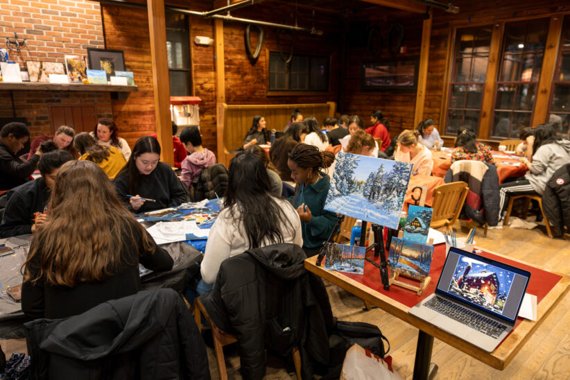 A table displays a painting and a laptop image of a different painting while students sit at tables around the Barn working on their own renditions of the same image