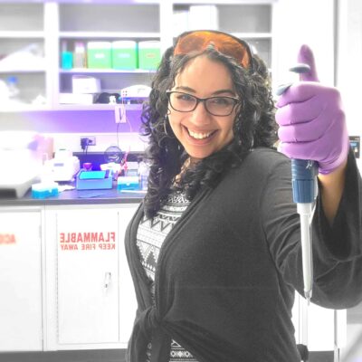 Katie Randolph stands in a lab with a gloved hand holding a scientific instrument and safety glasses perched on her head